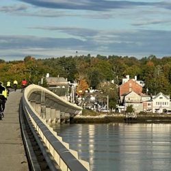 Bikers cross a bridge into Wicasset, Maine, with white church steeples poking above the trees