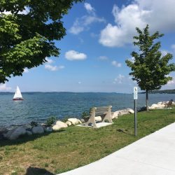 A bike path borders the water's edge, where a sailboat can be seen sailing in the distance.