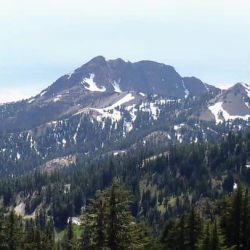 A young girl leans against a large rock with snow dotted Lassen Peak in the background